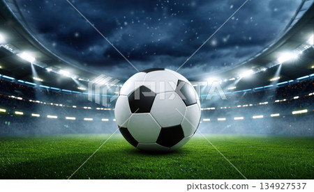 Soccer ball sits in the center of the field at a stadium during a night match under the lights and overcast sky Soccer ball sits in the center of the field at a stadium during a night match under the lights and overcast sky 134927537
