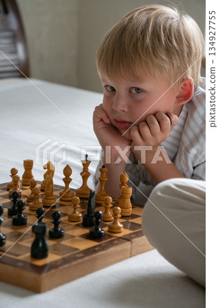 Young boy rests his chin on his hands while studying chessboard at home, showing focus, curiosity, thoughtful mood in soft natural light Young boy rests his chin on his hands while studying chessboard at home, showing focus, curiosity, thoughtful mood in soft natural light 134927755