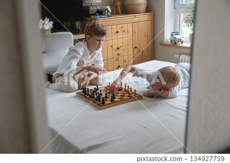 Two brothers sit on bed playing chess at home, focusing on board with thoughtful expressions in soft natural daylight Two brothers sit on bed playing chess at home, focusing on board with thoughtful expressions in soft natural daylight 134927759
