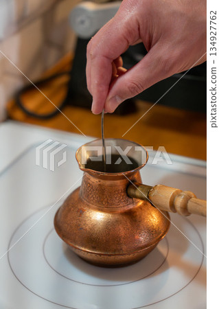 Hand stirs coffee in a copper cezve on a stovetop, showing a traditional brewing process with warm tones and rich texture 134927762
