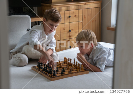 Two brothers sit on bed playing chess at home, focusing on board with thoughtful expressions in soft natural daylight Two brothers sit on bed playing chess at home, focusing on board with thoughtful expressions in soft natural daylight 134927766