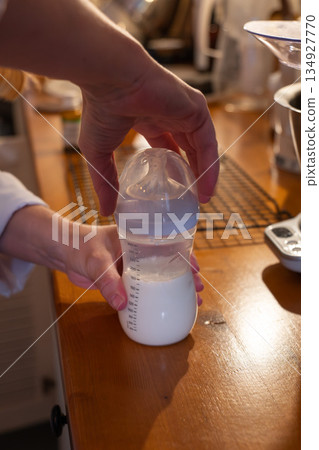 Hands prepare a baby bottle with milk on a wooden kitchen counter, showing careful pouring and a calm everyday parenting routine in warm light 134927770