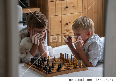Two brothers sit on bed playing chess at home, focusing on board with thoughtful expressions in soft natural daylight Two brothers sit on bed playing chess at home, focusing on board with thoughtful expressions in soft natural daylight 134927775