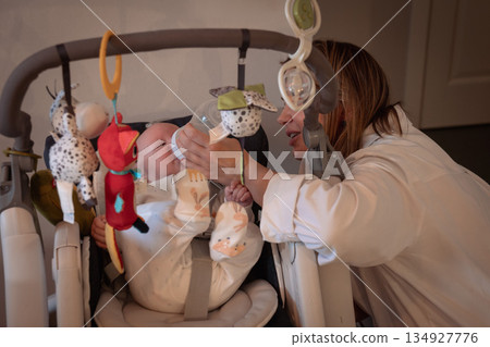 Mother gently interacts with her baby in home baby swing, surrounded by soft toys and warm indoor light, creating a caring and intimate family moment Mother gently interacts with her baby in home baby swing, surrounded by soft toys and warm indoor light, creating a caring and intimate family moment 134927776