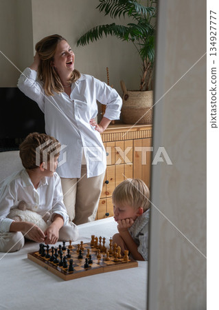 Mother stands beside her children playing chess on a bed, creating a warm family moment with soft daylight, neutral tones, and a calm home atmosphere Mother stands beside her children playing chess on a bed, creating a warm family moment with soft daylight, neutral tones, and a calm home atmosphere 134927777