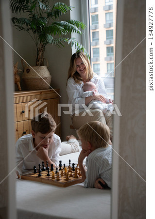 Mother sits by window holding baby while children play chess nearby, creating warm family moment with soft daylight and calm home atmosphere Mother sits by window holding baby while children play chess nearby, creating warm family moment with soft daylight and calm home atmosphere 134927778