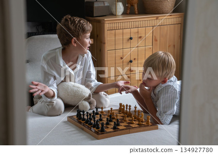 Two boys sit on bed playing chess at home, sharing a thoughtful moment with soft daylight, neutral tones, and a calm family atmosphere 134927780