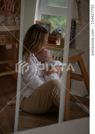Mother holding baby is seen as soft reflection in mirror near window, creating intimate family moment with gentle daylight, calm home mood Mother holding baby is seen as soft reflection in mirror near window, creating intimate family moment with gentle daylight, calm home mood 134927782
