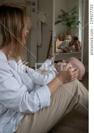 Mother sits on floor playing with baby while boys playing chess appear reflected in mirror, creating layered family moment in soft natural light Mother sits on floor playing with baby while boys playing chess appear reflected in mirror, creating layered family moment in soft natural light 134927783