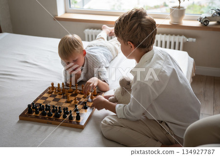 Two brothers sit on bed playing chess at home, focusing on board with thoughtful expressions in soft natural daylight Two brothers sit on bed playing chess at home, focusing on board with thoughtful expressions in soft natural daylight 134927787
