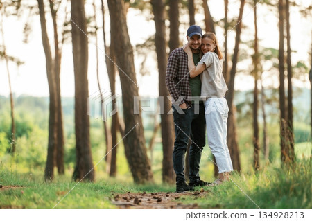 Standing and posing. Lovely couple are together in the forest at summer daytime Standing and posing. Lovely couple are together in the forest at summer daytime 134928213