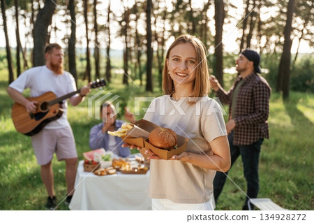 Woman is holding burger. Group of friends are having picnic on the field with food in eco boxes Woman is holding burger. Group of friends are having picnic on the field with food in eco boxes 134928272