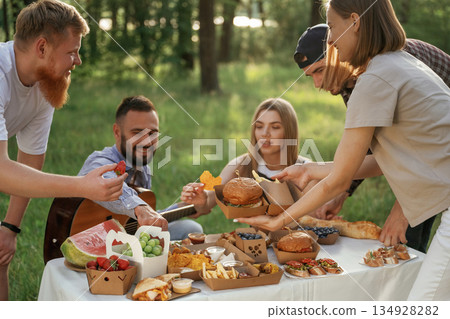 Sitting by the table. Group of friends are having picnic on the field with food in eco boxes 134928282