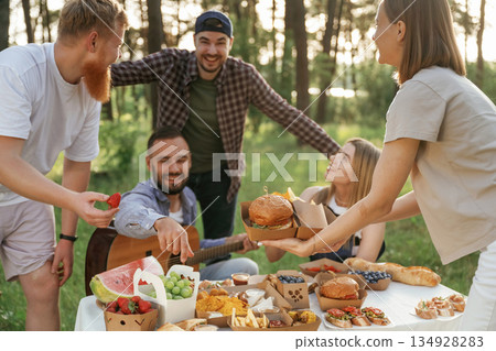 Guy is with acoustic guitar, having fun. Group of friends are having picnic on the field with food in eco boxes 134928283
