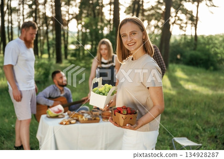 Strawberry in hands of woman. Group of friends are having picnic on the field with food in eco boxes Strawberry in hands of woman. Group of friends are having picnic on the field with food in eco boxes 134928287