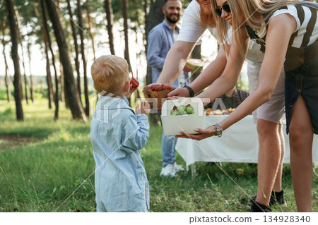 Cute little boy is standing with people. Group of friends are having picnic on the field with food in eco boxes Cute little boy is standing with people. Group of friends are having picnic on the field with food in eco boxes 134928340