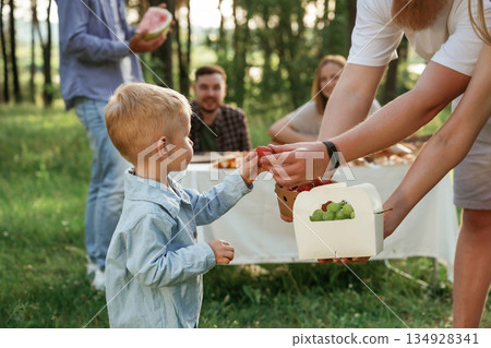 Cute little boy is standing with people. Group of friends are having picnic on the field with food in eco boxes Cute little boy is standing with people. Group of friends are having picnic on the field with food in eco boxes 134928341