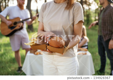 Woman is holding burger. Group of friends are having picnic on the field with food in eco boxes Woman is holding burger. Group of friends are having picnic on the field with food in eco boxes 134928367