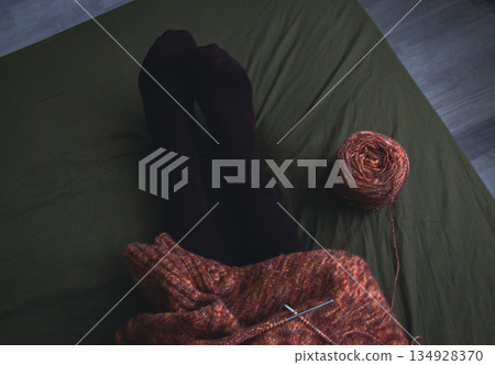 Melange brown sweater with knitting needles and women legs in velvet socks on dark green background. Knitting needles and balls of warm melange yarn. Girl legs and knitting work in process.  134928370