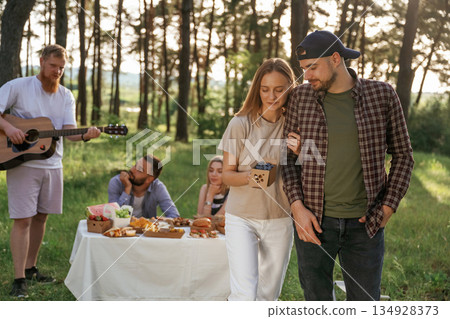 Beautiful couple is standing. Group of friends are having picnic on the field with food in eco boxes Beautiful couple is standing. Group of friends are having picnic on the field with food in eco boxes 134928373
