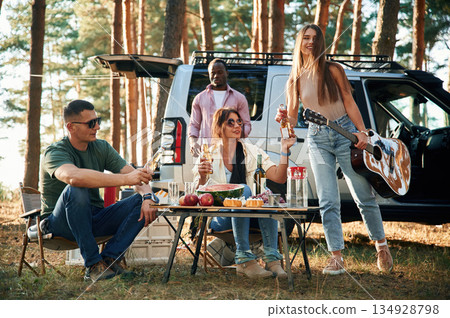 Sitting by the table with food, against car. Group of friends are together in the forest Sitting by the table with food, against car. Group of friends are together in the forest 134928798