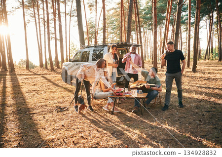 Drinking, eating, playing with dog. Group of friends are together in the forest Drinking, eating, playing with dog. Group of friends are together in the forest 134928832