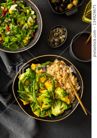 Healthy breakfast consists of a quinoa bowl with boiled eggs, green beans, and broccoli placed on a dark surface next to a cup of coffee and a woven cloth 134929675