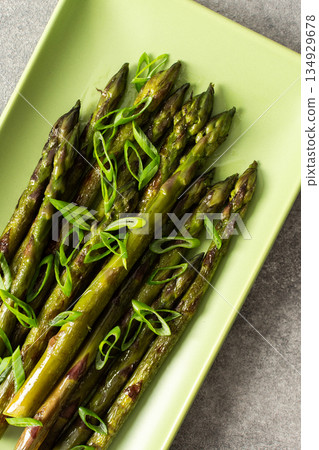 Asparagus spears are arranged neatly on a green plate, garnished with sliced green onions. This dish represents a simple yet flavorful vegetable choice for any meal 134929678
