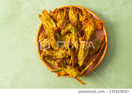 Golden-brown deep-fried zucchini flowers are arranged on a vibrant plate. The dish is set on a floral-patterned napkin, enhancing its appetizing appearance 134929683