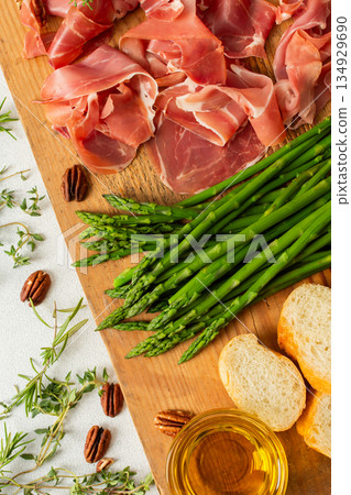 A beautifully arranged charcuterie board featuring slices of ham, blocks of cheese, fresh asparagus, and crunchy nuts. Perfect for enjoying with friends and family during special moments A beautifully arranged charcuterie board featuring slices of ham, blocks of cheese, fresh asparagus, and crunchy nuts. Perfect for enjoying with friends and family during special moments 134929690