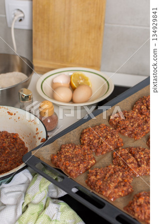 Liver cutlets are shaped and placed on a baking sheet. Ingredients like eggs, flour, and lemon sit on the counter 134929841
