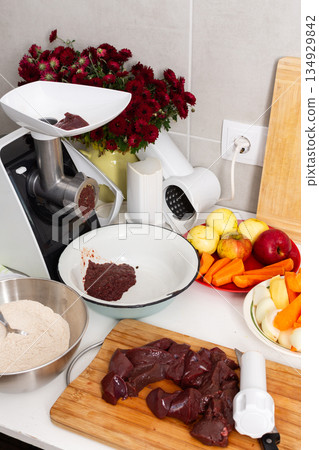 A woman is making liver cutlets in a city kitchen. She is using a grinder to prepare the meat. The kitchen has flowers on the balcony and autumn colors outside 134929842