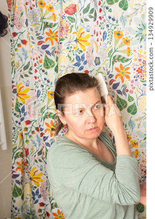 In a bright room decorated with a colorful floral curtain, a 50-year-old woman calmly brushes her hair as part of her morning routine, showcasing a peaceful start to her day In a bright room decorated with a colorful floral curtain, a 50-year-old woman calmly brushes her hair as part of her morning routine, showcasing a peaceful start to her day 134929909