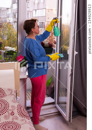 A 50-year-old woman dressed in colorful clothing cleans the windows on her balcony, getting her home ready for the winter. Flowers are placed around her, adding life to the scene 134929913