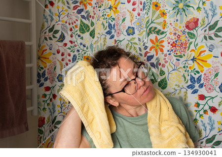 A 50-year-old woman starts her morning routine in the bathroom. She looks at herself in the mirror while holding a towel over her shoulder. The colorful background adds brightness A 50-year-old woman starts her morning routine in the bathroom. She looks at herself in the mirror while holding a towel over her shoulder. The colorful background adds brightness 134930041