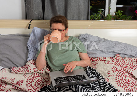 A 50-year-old woman sits on her bed using a laptop and holding a warm drink. She appears focused on her screen while surrounded by cozy blankets in her bedroom 134930043