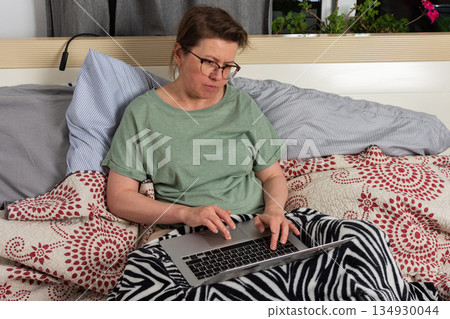A woman sits on bed in bedroom and works on laptop. She has blankets around her and seems focused on task while enjoying her own space 134930044