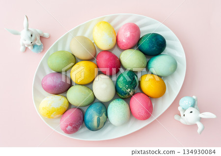 Multi-colored Easter eggs in an oval dish with decorative bunnies on a pink background, Happy Easter holiday Multi-colored Easter eggs in an oval dish with decorative bunnies on a pink background, Happy Easter holiday 134930084