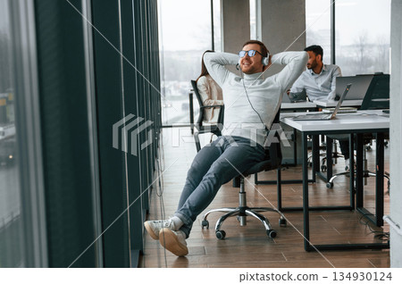 Relaxed, on the chair, in headphones. Men and woman are working in the office together Relaxed, on the chair, in headphones. Men and woman are working in the office together 134930124