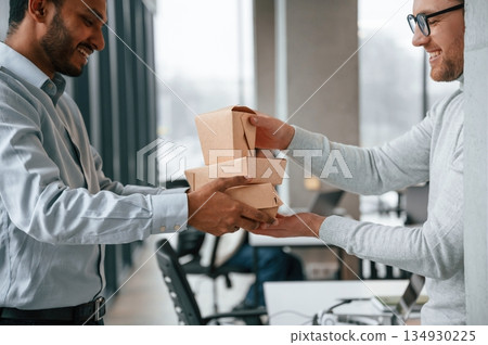 View from the side. Holding paper eco boxes. Two men are working in the modern office together View from the side. Holding paper eco boxes. Two men are working in the modern office together 134930225