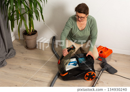 In a living room, a person is getting a vacuum cleaner ready for cleaning. The person is seated on the floor with the vacuum cleaner open and looks focused on the task In a living room, a person is getting a vacuum cleaner ready for cleaning. The person is seated on the floor with the vacuum cleaner open and looks focused on the task 134930240