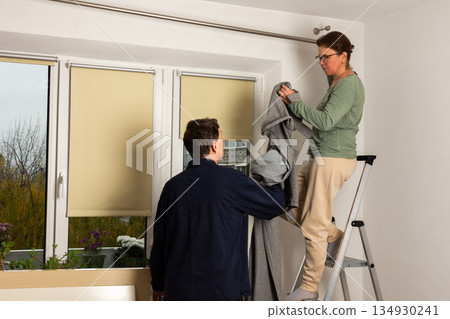 Married couple work together to hang freshly cleaned curtains in their living room after washing them. They are using a ladder to reach the curtain rod Married couple work together to hang freshly cleaned curtains in their living room after washing them. They are using a ladder to reach the curtain rod 134930241