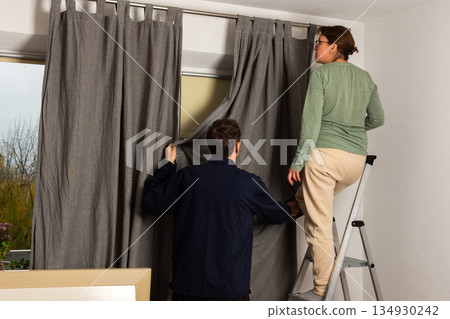 A married couple works together to hang freshly washed curtains in their living room. They position the curtains as they prepare for the autumn season in their home A married couple works together to hang freshly washed curtains in their living room. They position the curtains as they prepare for the autumn season in their home 134930242