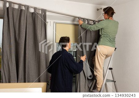 A married couple works together to hang freshly washed curtains in their living room. They position the curtains as they prepare for the autumn season in their home 134930243