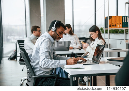 Man is sitting by laptop in headphones. Four people are working in the office together Man is sitting by laptop in headphones. Four people are working in the office together 134930307