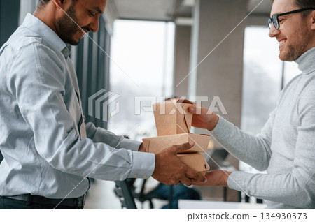 View from the side. Holding paper eco boxes. Two men are working in the modern office together View from the side. Holding paper eco boxes. Two men are working in the modern office together 134930373