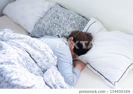 A middle-aged woman is sleeping soundly in her bed, wearing a sleep mask. She is surrounded by soft pillows and a warm blanket, taking a break from her daily routine 134930389