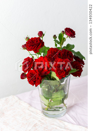 A vibrant bouquet of red roses is arranged in a clear glass vase, showcasing their lush green leaves. The soft pink background adds a gentle touch to the floral display A vibrant bouquet of red roses is arranged in a clear glass vase, showcasing their lush green leaves. The soft pink background adds a gentle touch to the floral display 134930422