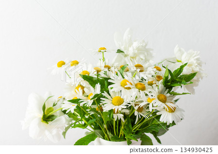 Fresh daisies are arranged in a decorative vase, brightening up a light room. The soft pink tablecloth enhances the cheerful atmosphere, showcasing spring flowers Fresh daisies are arranged in a decorative vase, brightening up a light room. The soft pink tablecloth enhances the cheerful atmosphere, showcasing spring flowers 134930425