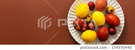 Vibrant Easter eggs in various colors rest on a white plate, showcasing shades of yellow, red, and speckled patterns, perfect for holiday decor and celebration, banner Vibrant Easter eggs in various colors rest on a white plate, showcasing shades of yellow, red, and speckled patterns, perfect for holiday decor and celebration, banner 134930448
